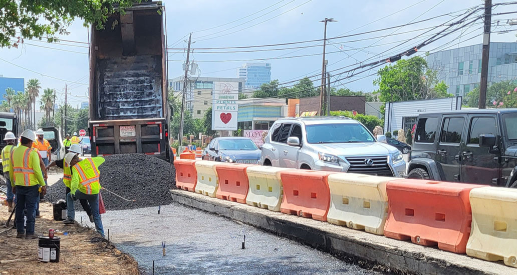 Dump truck unloading black asphalt base while construction workers spread it evenly on road on the corner of Westheimer and Weslayne.