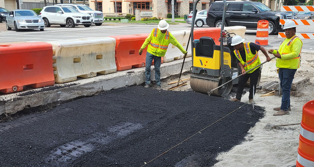 Construction workers spreading and use a steel roller to flatten the freshly placed black asphalt surface on the corner of Westheimer and Weslayne.