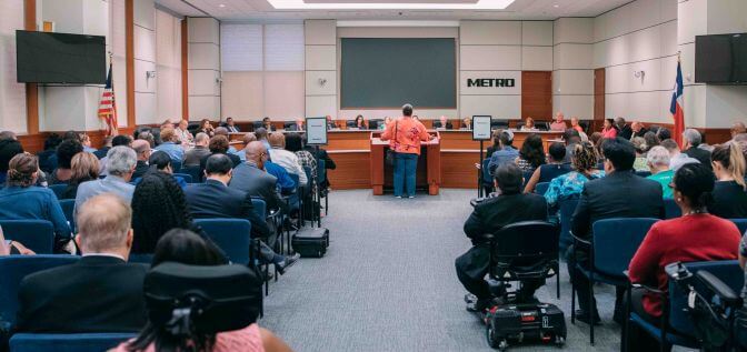 View from the back of the METRO boardroom during a meeting of the Board of Directors with many people in attendance including some in motorized wheelchairs.