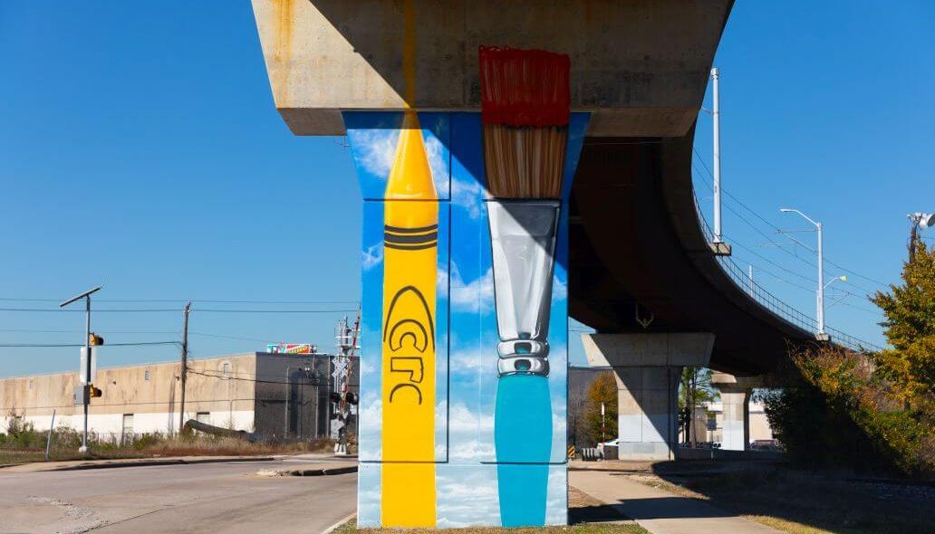 Mural depicting a yellow crayon next to an artist's brush on a vertical column under METRORail tracks near Roosevelt Elementary School at 6700 Fulton St. in Houston