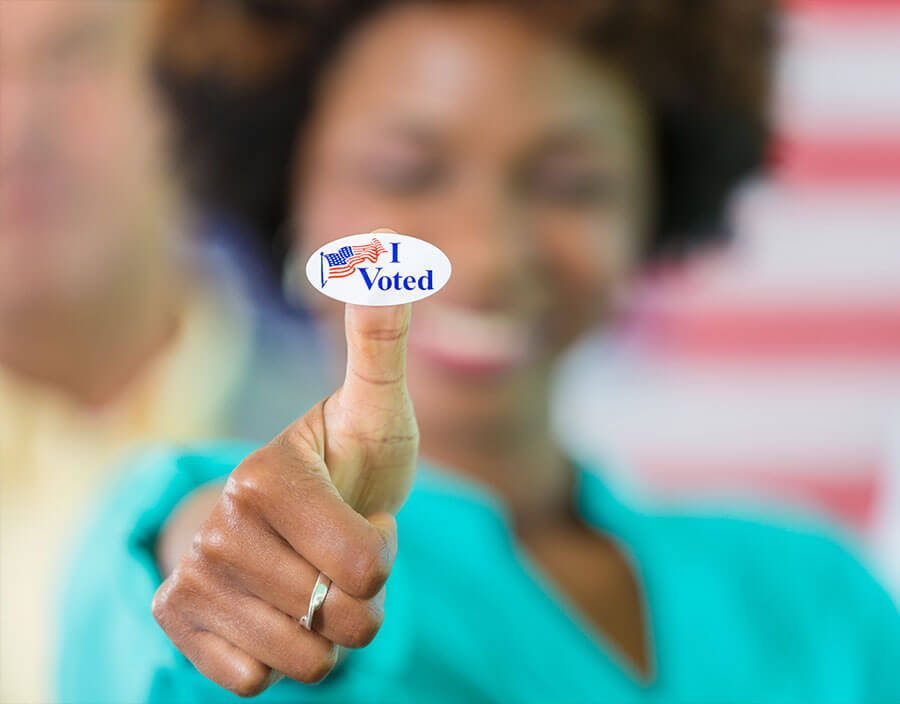 A woman holding an "I Voted" sticker towards you in the foreground on her thumb finger.