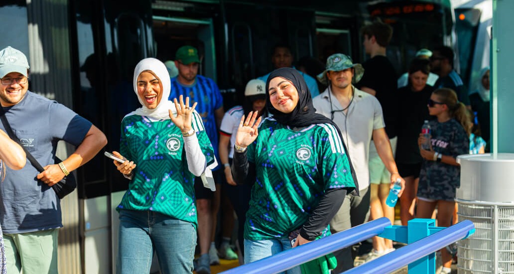 Two riders wearing Saudi Arabia soccer jerseys waving while exiting the train.