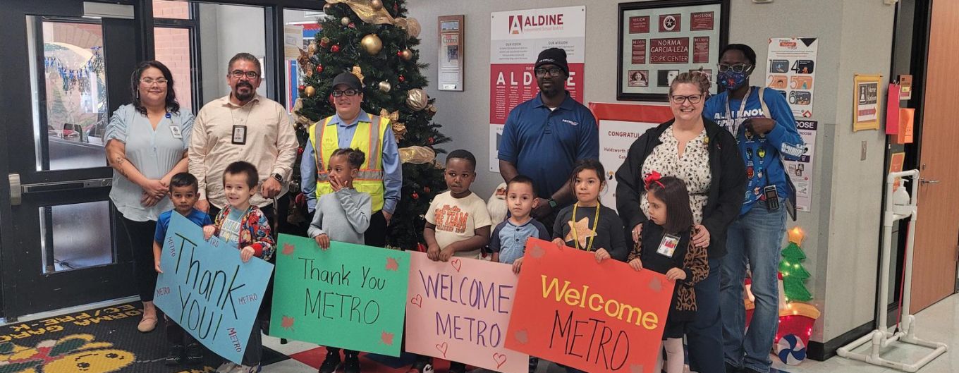 Young children hold signs in front of a Christmas tree thanking and welcoming METRO employees.