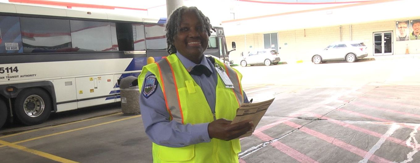 Female bus operator with yellow safety vest stands in front of a bus holding a checklist and smiling.