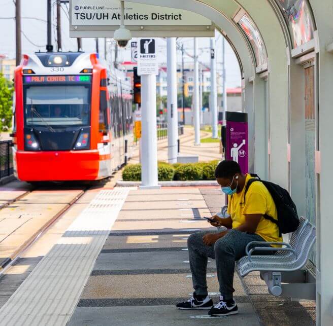 4845-Customer-Seated-at-METRORail-Platform-656X643 Customer seated on a bench and looking down at their phone on a METRORail platform as train approaches station