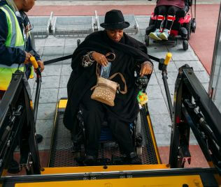 1098-Paratransit-Lift-316X268 Point of view photo of a rider in a wheelchair on an electric lift about to board a METROLift van.